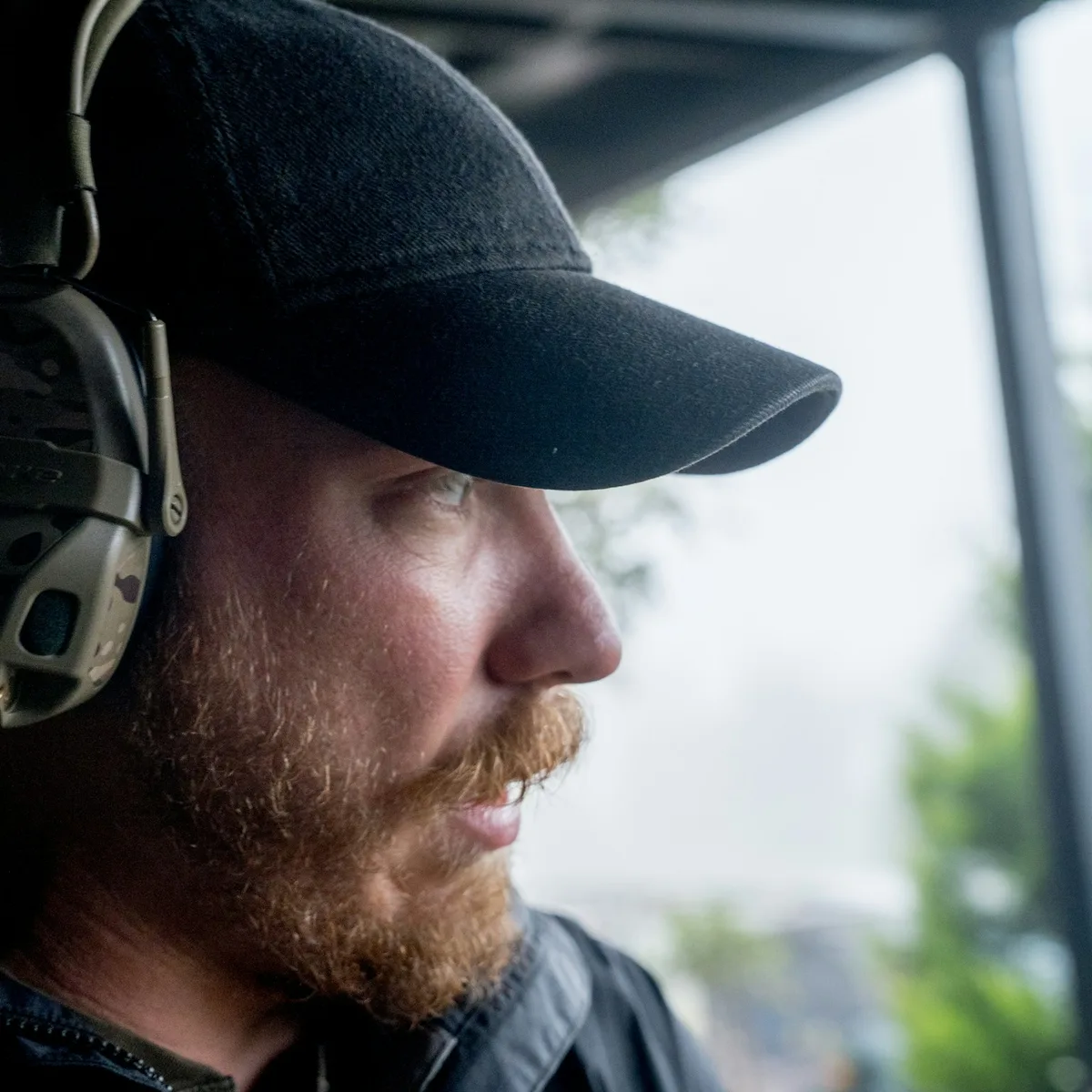 Picture of white man, age 25-35, medium build, strawberry blond beard, blue eyes. Wearing black hat, black rain jacket, multicam hearing protection.